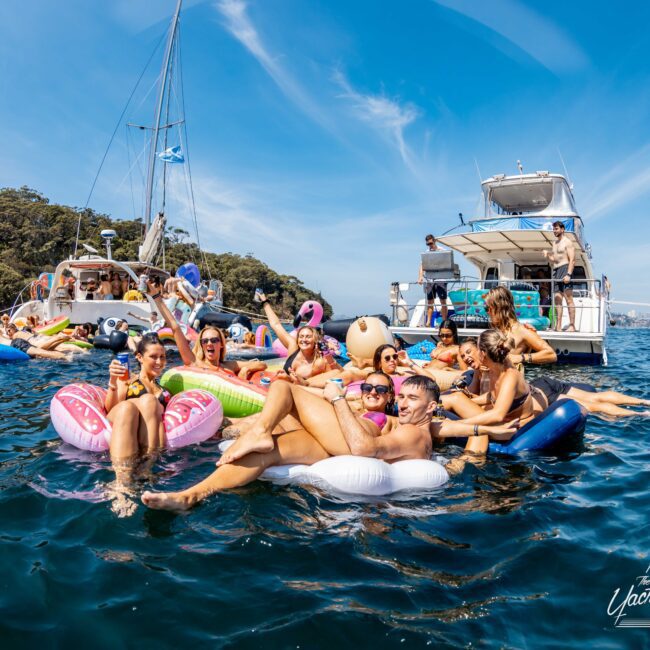 A group of people are relaxing on colorful inflatables in a sunny, blue ocean. Two boats are anchored nearby, and several others are enjoying the water. The atmosphere is lively and festive.