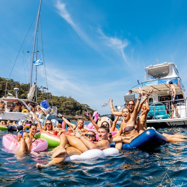 A group of people are having fun on inflatables in the water near a yacht and sailboat, on a sunny day. Some are raising their arms joyfully. The sky is clear and blue, and the boats are anchored nearby.
