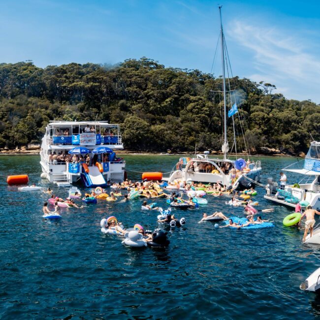 A lively scene on the water with people floating on inflatables and swimming around several anchored boats. The background features a tree-covered island under a bright blue sky. The gathering appears festive and joyful.
