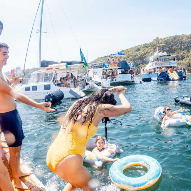 A woman in a yellow swimsuit splashes water playfully as she joins others on inflatable rings in the sea. Boats and yachts in the background with people enjoying the sunny day create a lively atmosphere.