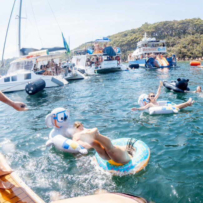 People enjoy a sunny day on the water, swimming with inflatable toys near several docked boats. The scene is lively, featuring a boat party with music. The backdrop includes trees and a clear sky.