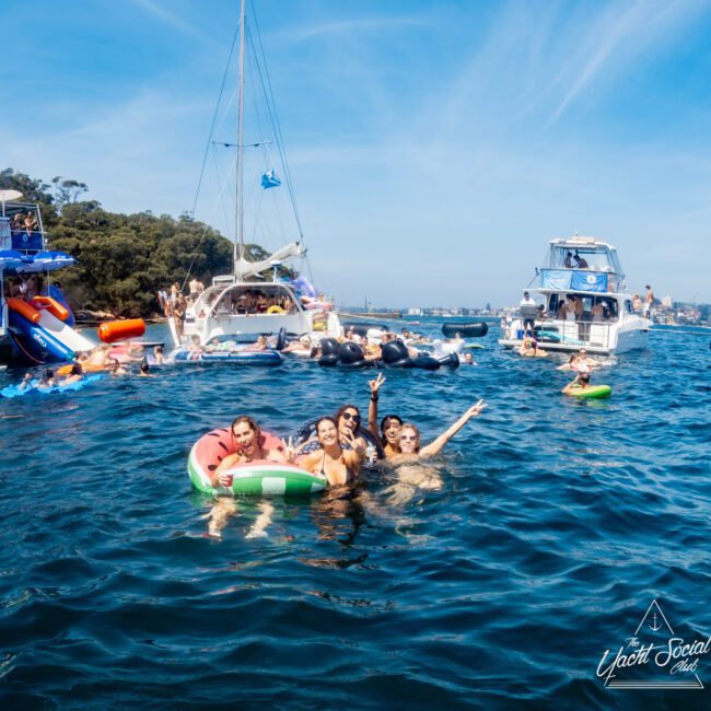 A group of people enjoying a sunny day on the water, surrounded by boats. Some are swimming, while others relax on inflatable rings. The scenery includes clear blue water and trees in the background. A logo in the corner reads "Yacht Social Club.