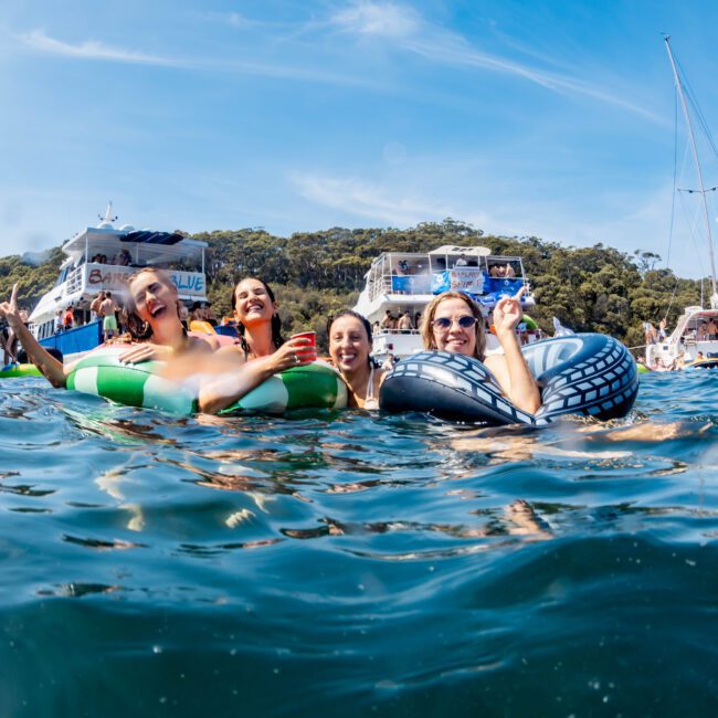 A group of people floating in water, smiling and holding drinks. They are using inflatable rings. Yachts are in the background, and the sky is clear with a few clouds.