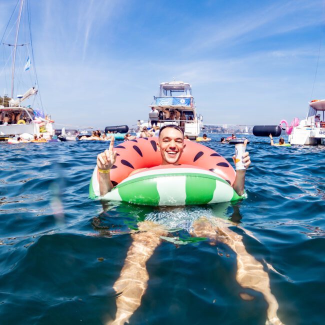 A person is floating in the water on a watermelon-themed inflatable ring. They are smiling, giving two thumbs up, surrounded by several boats in the background on a sunny day. The sky is clear with streaks of wispy clouds.