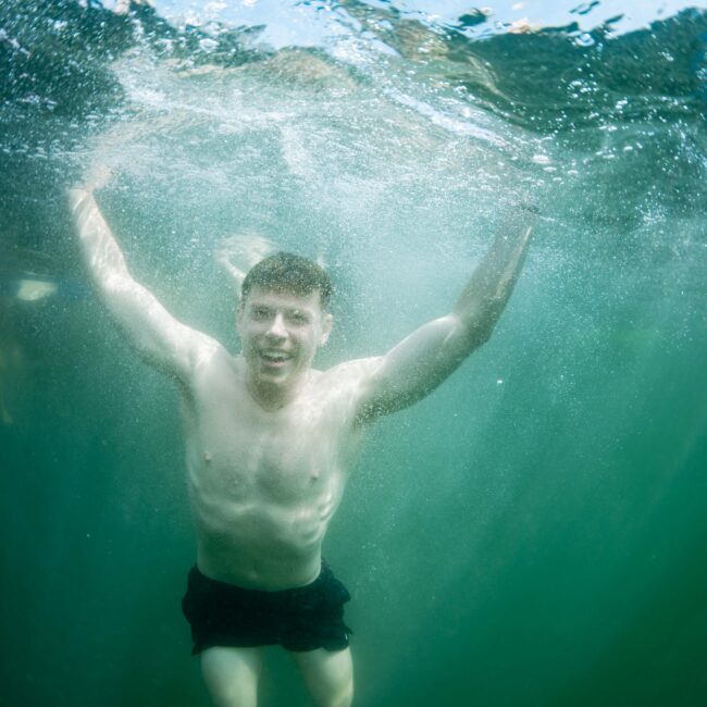 A person with short hair is swimming underwater. They are wearing black swim trunks and appear to be smiling, with arms outstretched. The water is clear, and the surface is visible above.