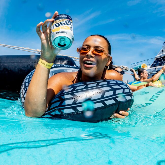 A person with sunglasses relaxes in a pool float, holding up a can of Suntory 196 Double Lemon. The sky is clear and blue, with boats and other people enjoying the water in the background.