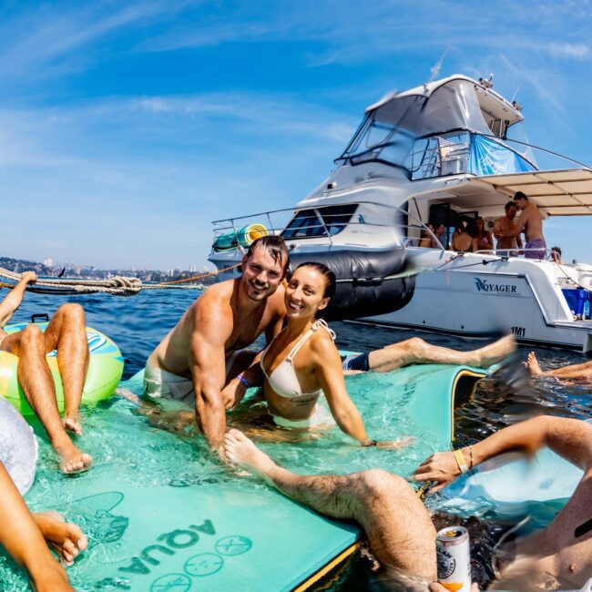 A man and woman smiling on a floating mat in the water, surrounded by others. There's a yacht in the background with people on board. The scene is lively and sunny, suggesting a fun day at sea.