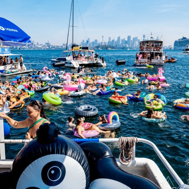A lively scene of people enjoying a sunny day in the water with colorful inflatable pool floats, including a panda, unicorn, and tire. Boats are nearby, and a city skyline is visible in the background under a clear blue sky.