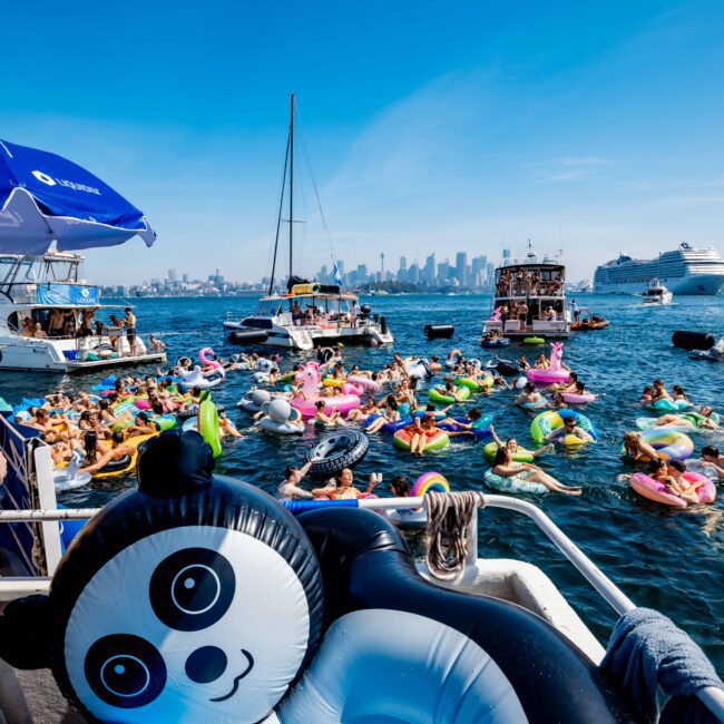 A lively scene with people on colorful inflatable floats in the sea near several boats, including a cruise ship in the background. A large panda float is in the foreground under a bright blue sky. City skyline visible in the distance.