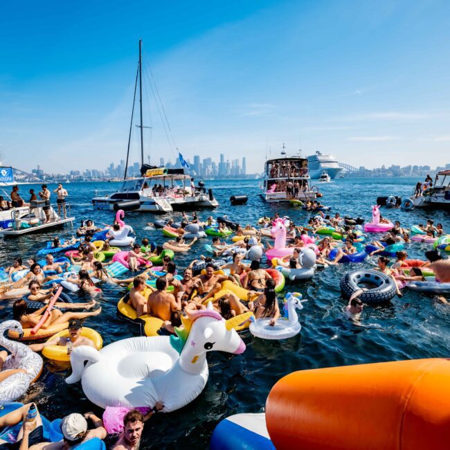 A lively boat party on a sunny day with people swimming and lounging on colorful inflatable floats in various shapes, such as swans and flamingos, near several yachts. A city skyline is visible in the background.
