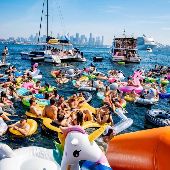People are enjoying a sunny day on the water, floating on inflatable rafts and unicorn floats near several boats. The skyline of a city is visible in the background.