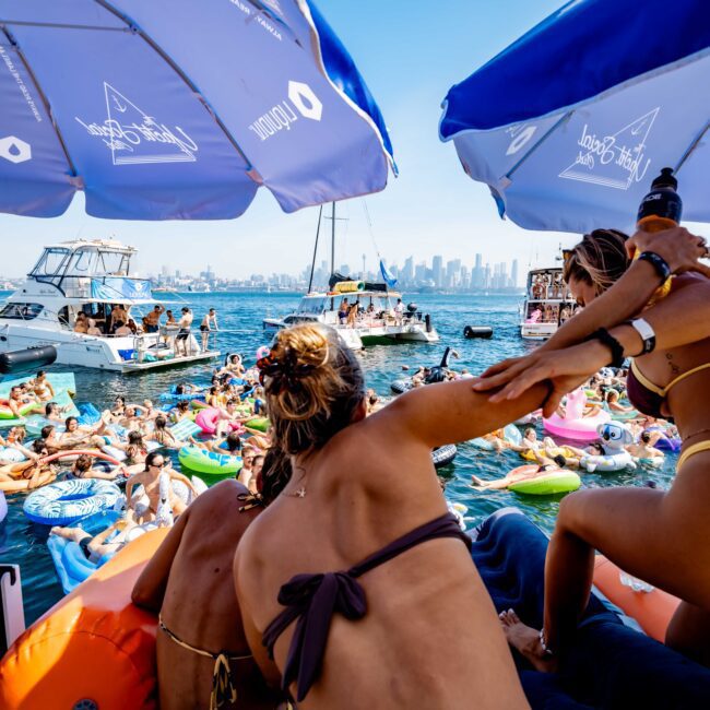 People enjoying a sunny day on the water under large blue umbrellas. Many are in swimsuits, lounging on inflatables near boats. The city skyline is visible in the distance.