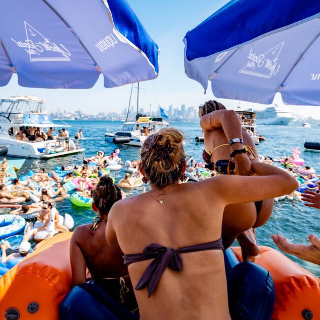 People on a boat party under umbrellas watch others in a crowded, colorful float-filled sea. The background features a city skyline, and someone is helping a child into the water. It's a vibrant, sunny day.