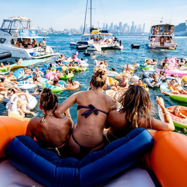 A lively scene of people on colorful inflatable rafts and floats gathered around several boats in a sunny harbor. The city skyline is visible in the background. Three people sit on an orange inflatable, facing the crowd.