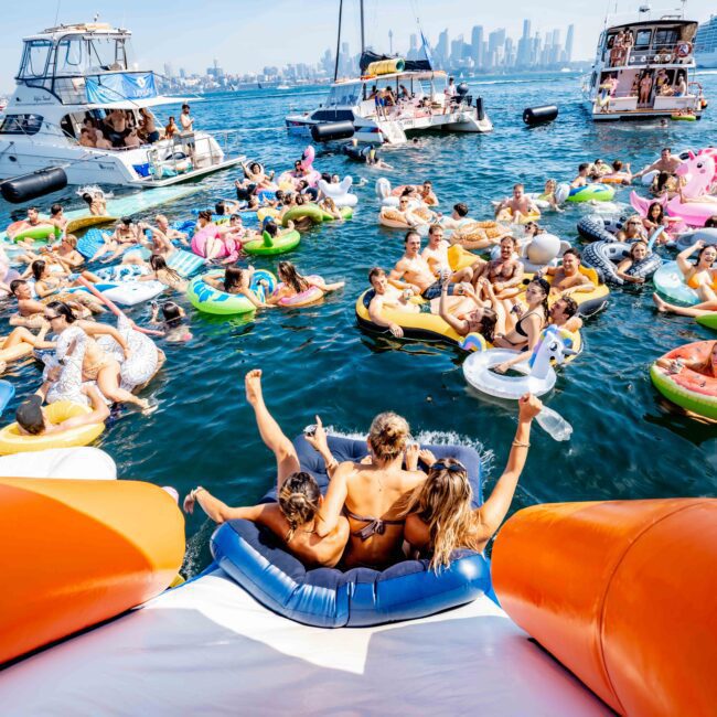 A lively scene of people relaxing on inflatable floaties in a sunny harbor. Boats and a cruise ship are visible in the background, with a distant city skyline. Some individuals hold drinks, enjoying the festive atmosphere.