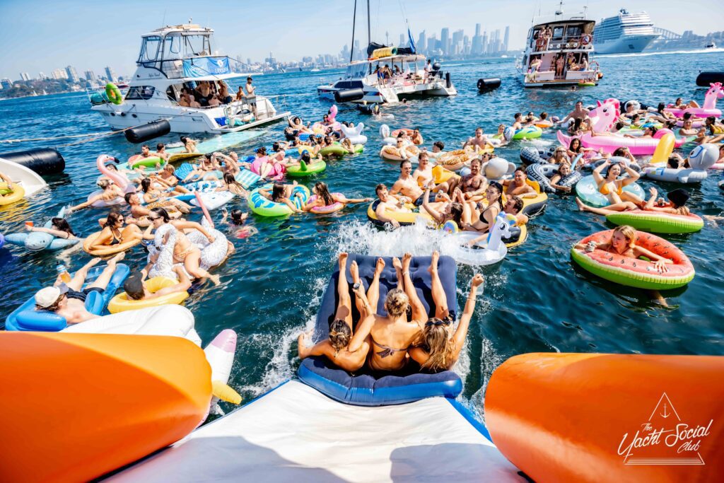 A lively gathering of people on colorful inflatable floats in a bay, some splashing down a slide from a boat. The background features several yachts and a city skyline, creating a festive, summer atmosphere.