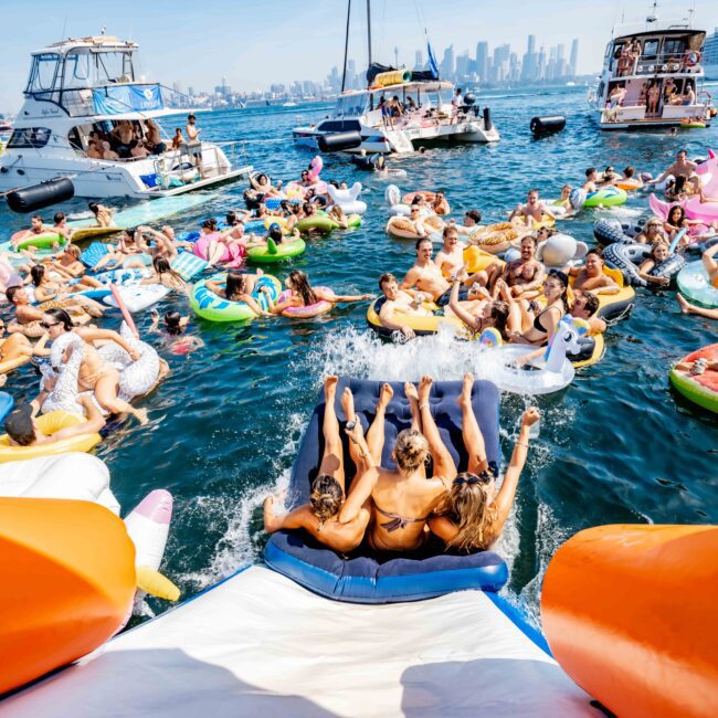 A lively gathering of people on colorful inflatable floats in a bay, some splashing down a slide from a boat. The background features several yachts and a city skyline, creating a festive, summer atmosphere.