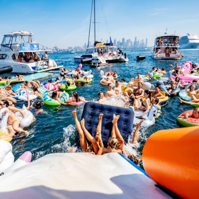 A lively summer scene with numerous people on inflatable floats in the water, surrounded by boats. The floats are colorful, and the city skyline is visible in the background under a clear blue sky. The atmosphere is festive and vibrant.