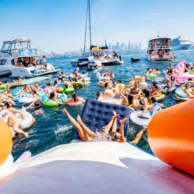 People on colorful inflatable floats enjoy a sunny day in the water near several boats. The city skyline is visible in the background. The scene is lively, with many participants relaxing and having fun.