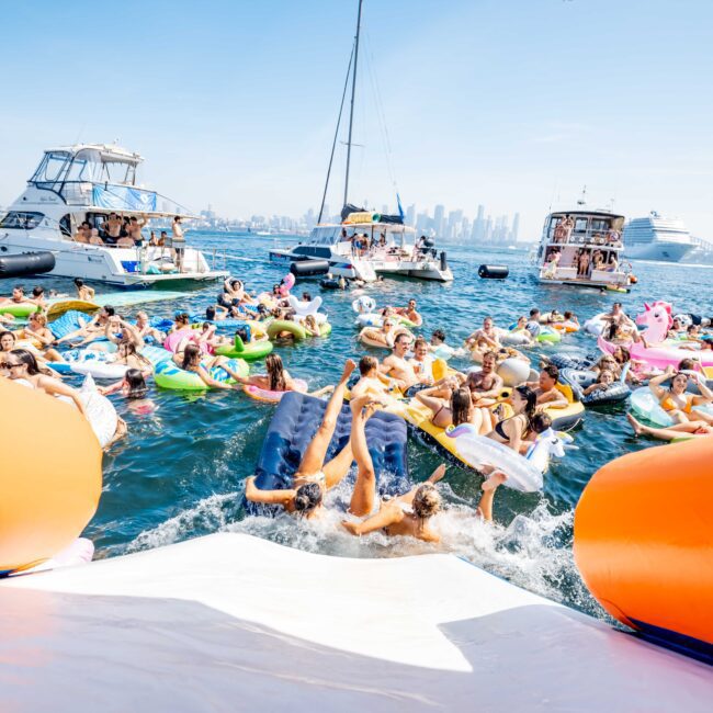 A large group of people relax on colorful inflatable floats in the water near several boats. The scene is lively under a bright blue sky, with a city skyline in the background. Orange slides are visible in the foreground.