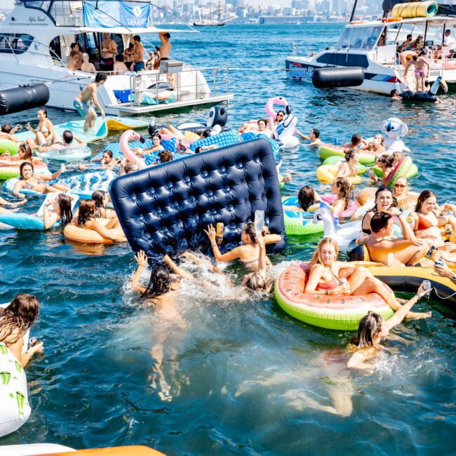A lively scene unfolds on the water with numerous people enjoying various colorful inflatable floats. Some are splashing around a large blue mattress float in a busy marina, set against a backdrop of boats and a distant city skyline.