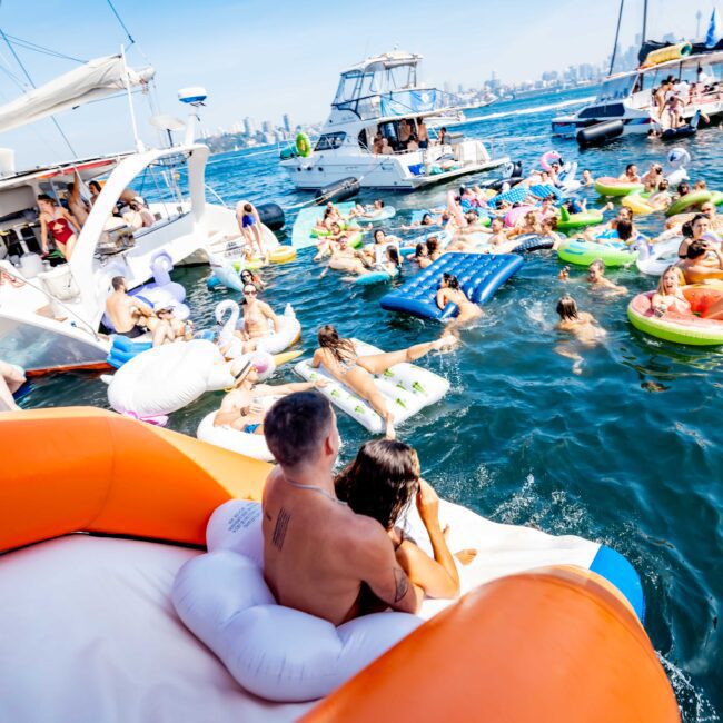 A lively scene on the water with people on a yacht and in colorful inflatable rafts. The skyline is visible in the background. Adults are enjoying a sunny day, swimming and relaxing.