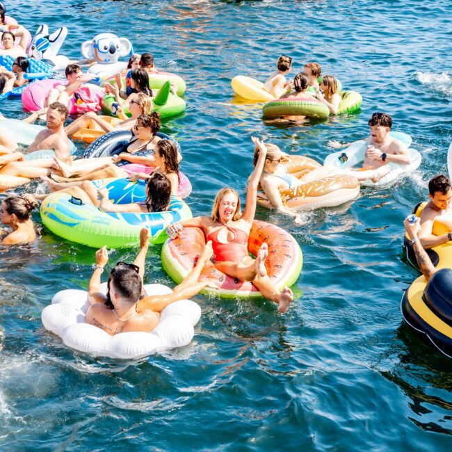 A group of people enjoying a sunny day on colorful inflatable floats in the water. A yacht is visible in the background. Smiles and relaxed postures suggest a fun, leisurely atmosphere.