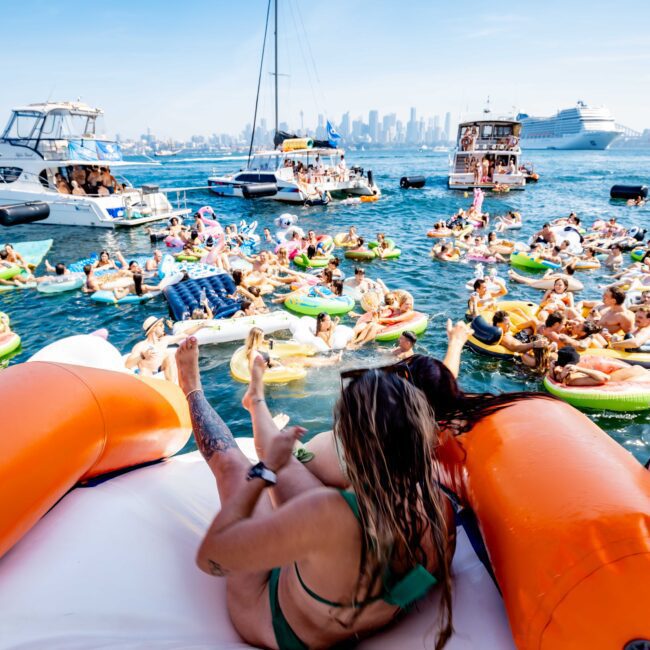 People relaxing on floats and boats in a vibrant harbor scene. The water is filled with colorful inflatable rafts, and the city skyline is visible in the background. A cruise ship and several yachts are anchored nearby. Sunny, festive atmosphere.