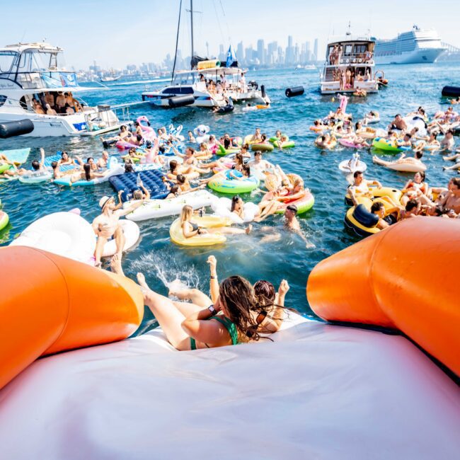 A vibrant scene of a boat party with people on colorful inflatables in the water. Boats and a cruise ship are in the background, along with a city skyline. The foreground shows people sliding into the water from an inflatable slide.