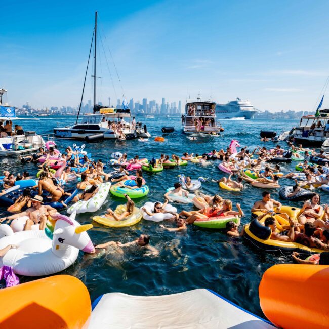 A large group of people relax on colorful inflatable floats in the water, surrounded by boats. The city skyline is visible in the background under a clear blue sky. Vibrant atmosphere with laughter and socializing.