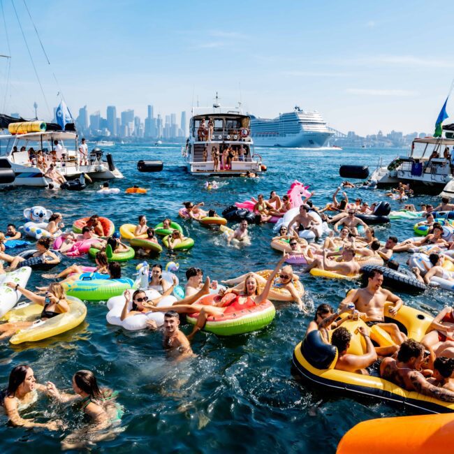 A lively scene of people enjoying a sunny day on inflatable pool floats in the water, surrounded by boats. The city skyline is visible in the background, and everyone appears to be having fun under clear blue skies.