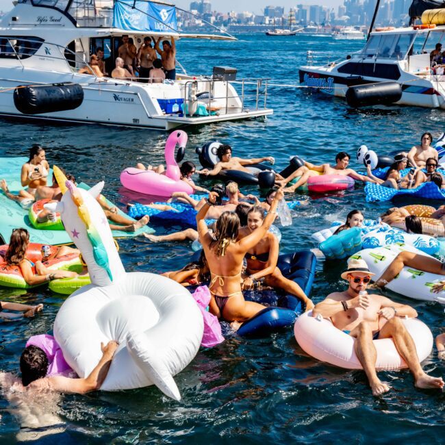A lively gathering of people on colorful inflatable pool floats in the water near several yachts. The city skyline is visible in the background. Participants are dancing, chatting, and enjoying the sunny day.
