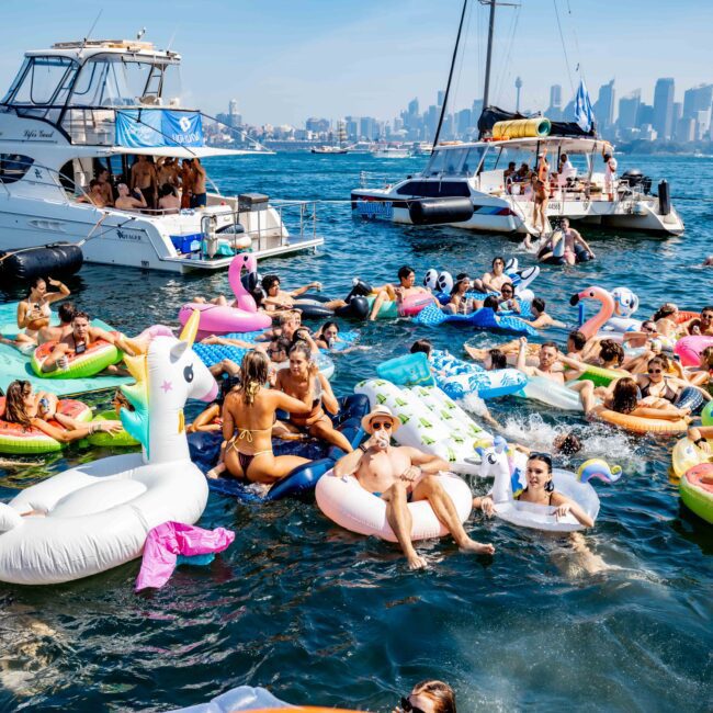 A lively scene on the water with people enjoying a sunny day on colorful inflatable pool floats, including unicorns and flamingos. Boats are anchored nearby, and a city skyline is visible in the background.