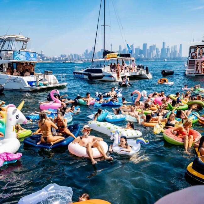 A lively scene of people lounging on colorful inflatable floats, including unicorns and flamingos, in a bustling harbor. Several boats float nearby under a clear blue sky, with a city skyline visible in the background.