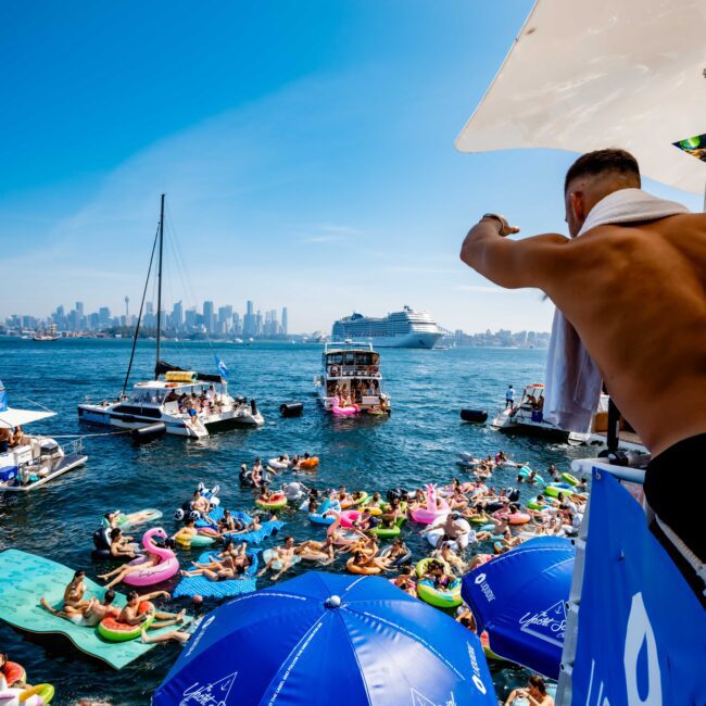 A lively scene on the water features numerous people on inflatable floats. Boats are anchored nearby, and a large cruise ship is in the distance. Blue umbrellas with a logo provide shade on the deck, and a person looks out from above.