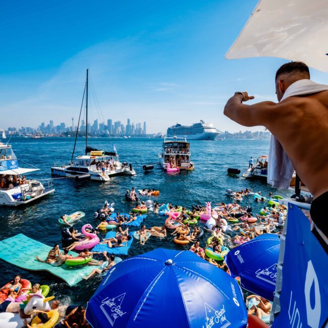 A lively boat party scene on a sunny day, with people on inflatable floats and boats in the water. The city skyline is visible in the background. A man on the deck watches the festivities, and blue umbrellas display the "Yacht Social Club" logo.