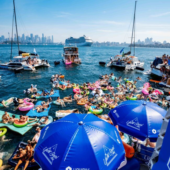 A bustling water scene with numerous people relaxing on colorful inflatable floats on a sunny day. Many boats surround them, and a cruise ship is in the distance. Bright blue umbrellas with logos provide shade. The skyline is visible in the background.
