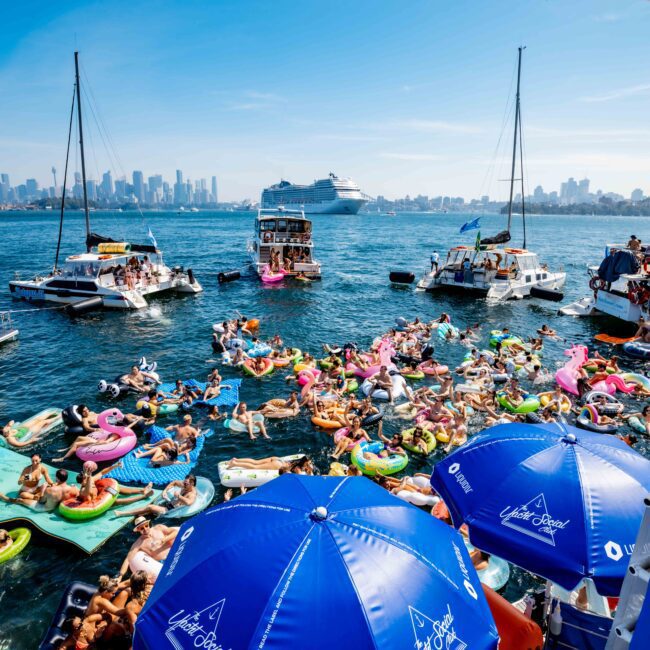 A crowded waterfront scene with numerous people on colorful inflatable floats and mats in the water. Several boats, including a large cruise ship, are in the background. Blue umbrellas with "Yacht Social Club" are visible in the foreground.