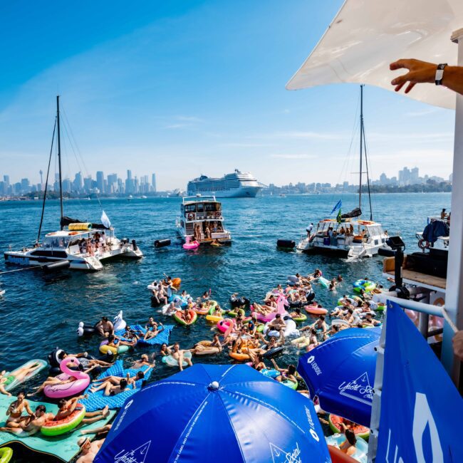 A lively boat party on a sunny day, with people on inflatables and yachts in the water. The city skyline and a cruise ship are visible in the background. Blue umbrellas with a logo are in the foreground, along with someone's arm gesturing.