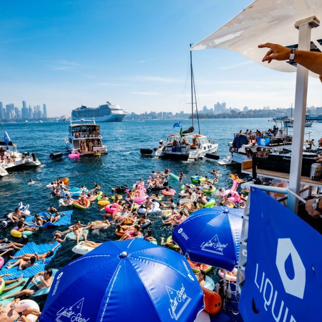 A lively scene on the water with people on inflatables and boats, enjoying a sunny day. The backdrop features a coastal city skyline and a cruise ship. A few individuals are on a boat pointing towards the distance, with blue umbrellas visible in the foreground.