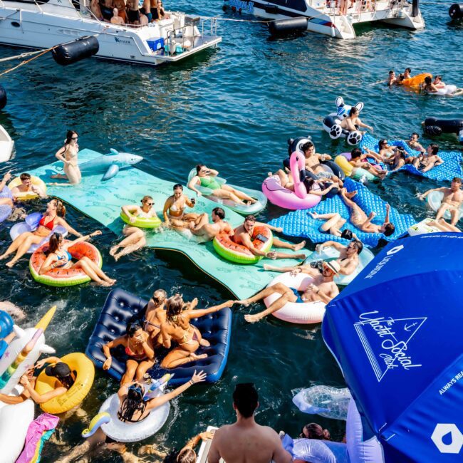 A lively scene on a sunny day with people enjoying a yacht party. Several yachts are anchored close together, and groups of people are relaxing on colorful inflatables in the water, with a city skyline visible in the background.