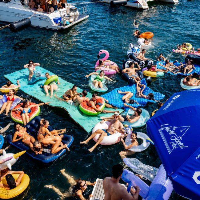 Aerial view of a festive scene with people on colorful inflatable floats in the water, surrounded by boats. Participants are enjoying the sunny day, socializing, and relaxing. A lively and cheerful atmosphere prevails.