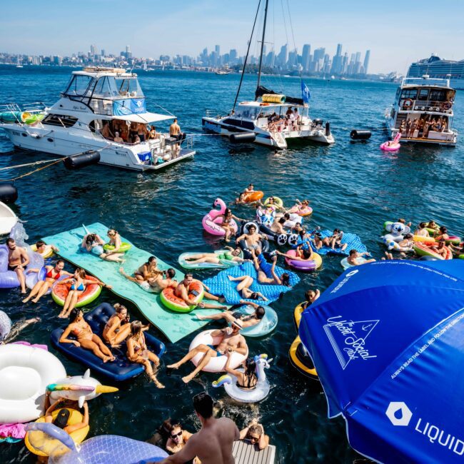 A lively scene on the water with numerous people on inflatable floats and a dock, surrounded by boats. The skyline of a city is visible in the background under a clear blue sky. Bright, colorful floats add to the festive atmosphere.