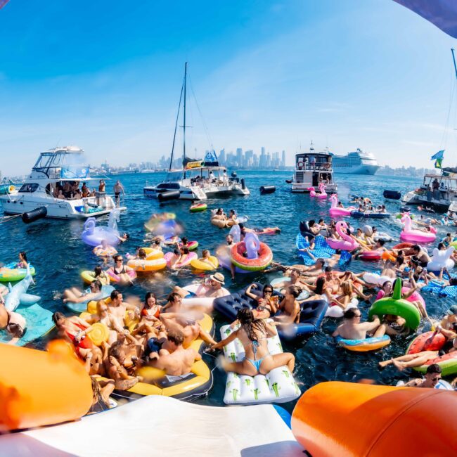 A festive scene on the water with numerous people floating on inflatable pool devices. Boats and yachts are anchored nearby with a city skyline in the background. The atmosphere is lively and colorful under a clear blue sky.