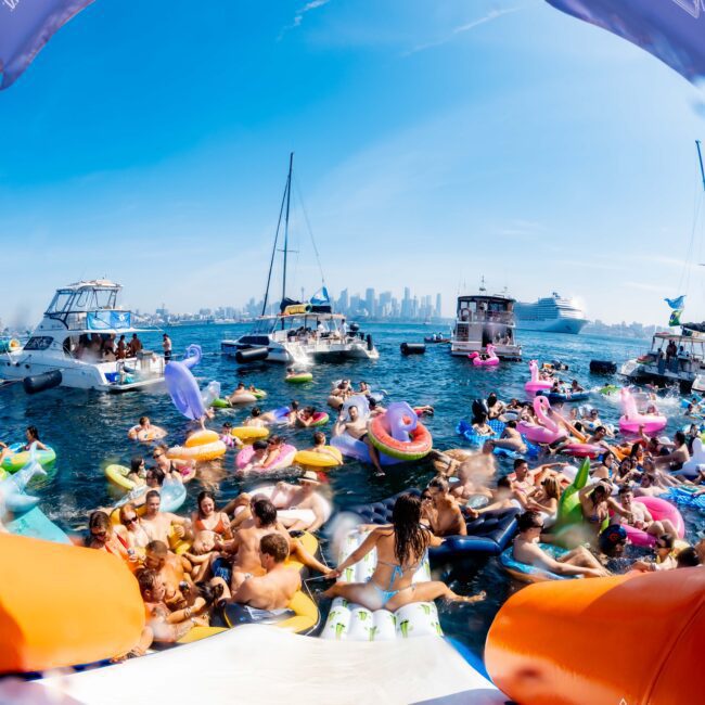 A lively boat party with numerous people on colorful inflatables in the water. Boats are anchored nearby, and a city skyline is visible in the background under a clear blue sky.