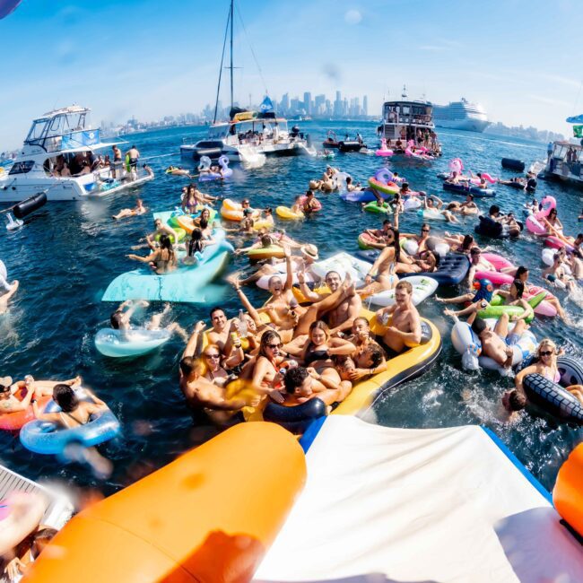 A lively waterfront party with people on colorful inflatable floats and boats in the water. The background shows a city skyline under a clear blue sky. Participants are enjoying the sunshine and festivities.
