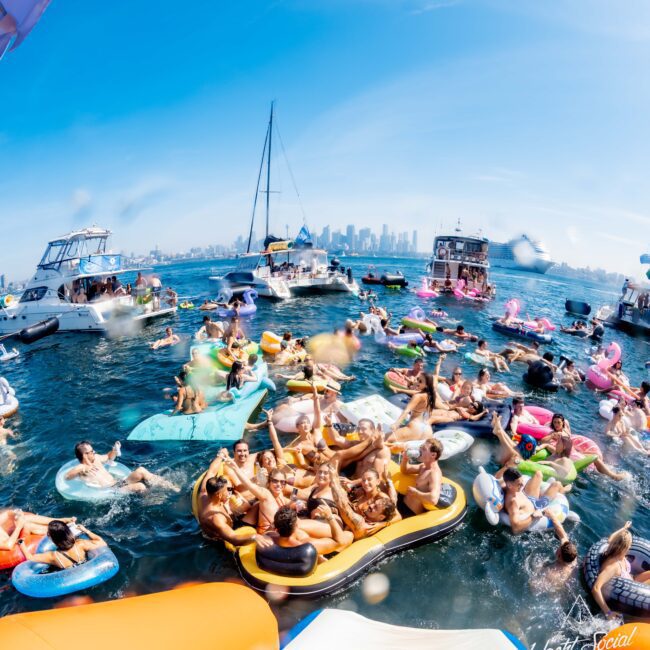 A lively group of people enjoy a sunny day on colorful inflatables in a large body of water. Several boats are anchored nearby. The background features a city skyline under a clear blue sky.