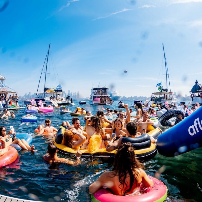 A lively scene of people enjoying a sunny day on the water. They are on colorful inflatables near several boats. The sky is clear, with visible city buildings in the background. The vibe is energetic and festive.
