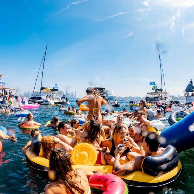 A lively scene on the water with numerous people enjoying themselves on inflatable rafts and floaties. Boats and yachts surround the group under a clear blue sky. The water is filled with colorful pool floats and cheerful partygoers.