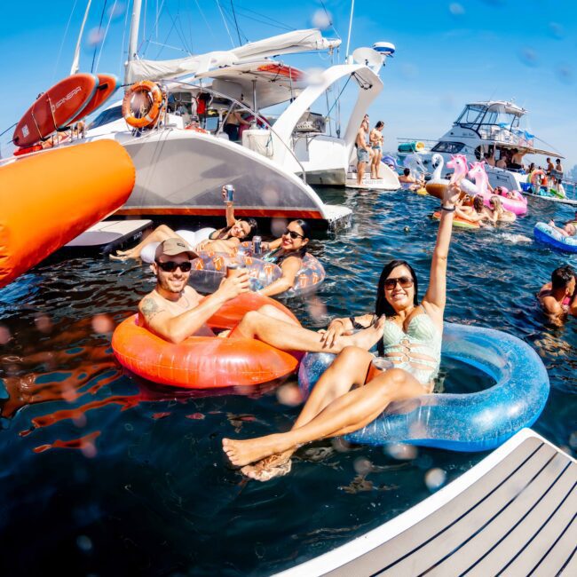 People are floating on inflatable tubes in a sunny, blue ocean with boats in the background. They are smiling, holding drinks, and enjoying the water. There are colorful inflatables, including a flamingo, adding to the festive atmosphere.